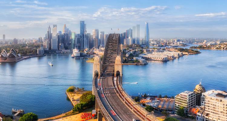 Aerial view of Sydney Harbour Bridge with city skyline and Opera House beside deep blue water.