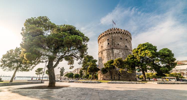 Historic White Tower of Thessaloniki rises behind shady pine trees on a sunny plaza.