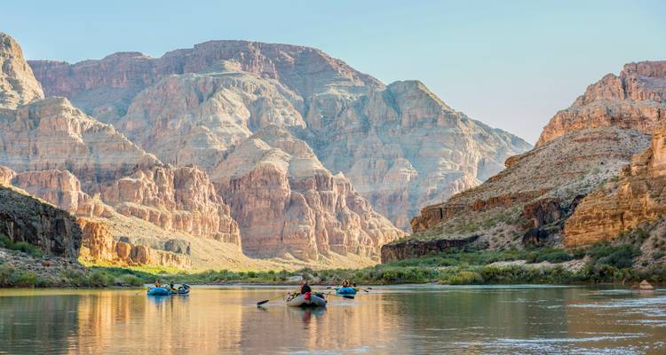 Rafters paddle along a calm river beneath towering canyon walls in warm light.