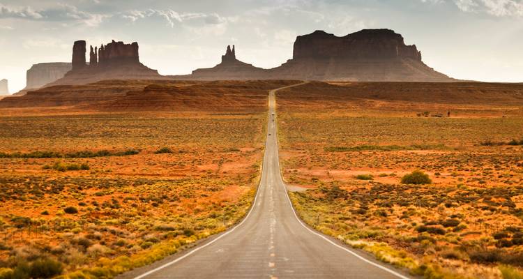A long straight highway leads toward Monument Valley’s dramatic mesas across open desert.