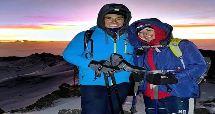 Two climbers bundled in winter gear smile at sunrise on a snowy mountain ridge