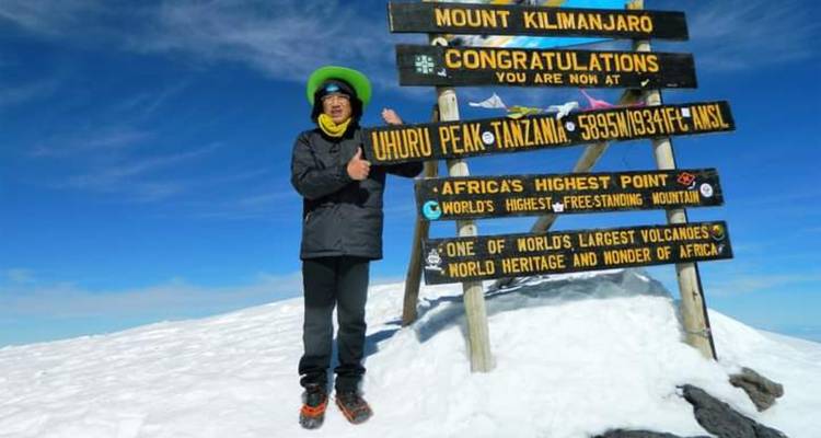 Solo climber gives thumbs up beside the Uhuru Peak sign on snowy Kilimanjaro summit