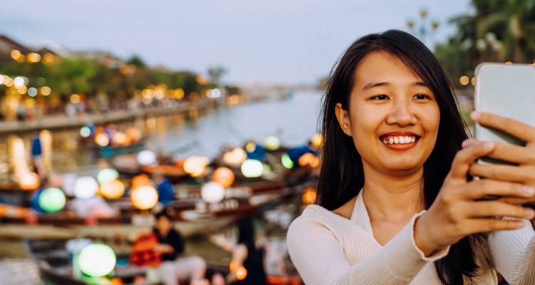 A smiling woman takes a selfie beside lantern-lit boats on a riverside evening in Hoi An.