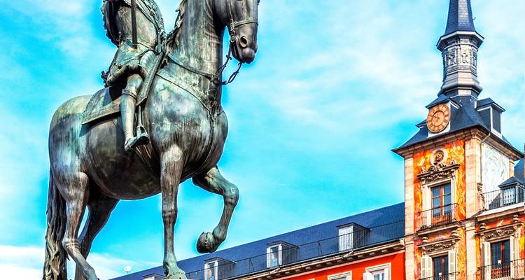 Close-up of bronze equestrian statue in Plaza Mayor with colorful painted façade and tower behind.