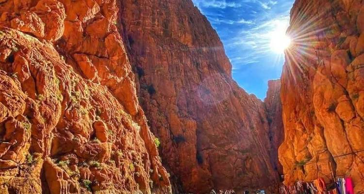 Sunburst over towering orange cliffs of Todra Gorge with tiny visitors walking along the canyon floor.