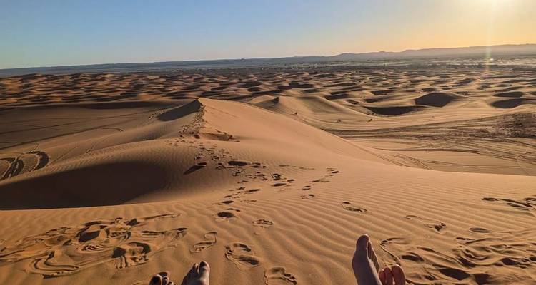 View from atop a giant sand dune with footprints and distant desert landscape at sunset.