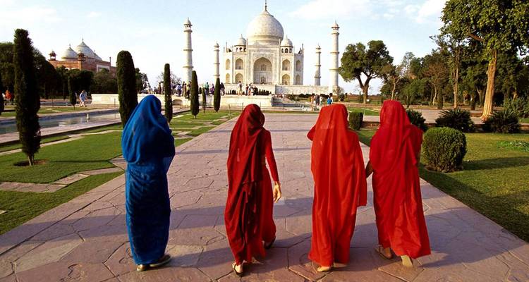 Four women in colourful saris walk toward the iconic white marble Taj Mahal across its garden path.