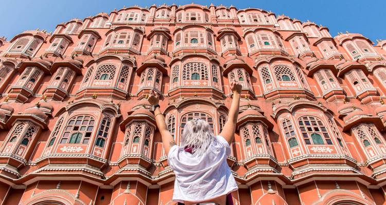 A traveller raises jubilant arms before Jaipur’s towering pink Hawa Mahal façade under a blue sky.