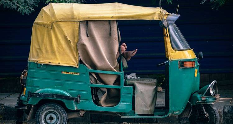 An auto-rickshaw driver relaxes with feet up inside a parked green and yellow tuk-tuk.