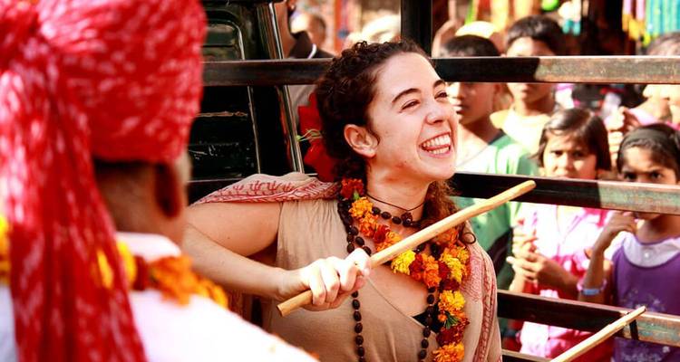 A joyful woman participates in a street festival, smiling and swinging a stick while locals watch.