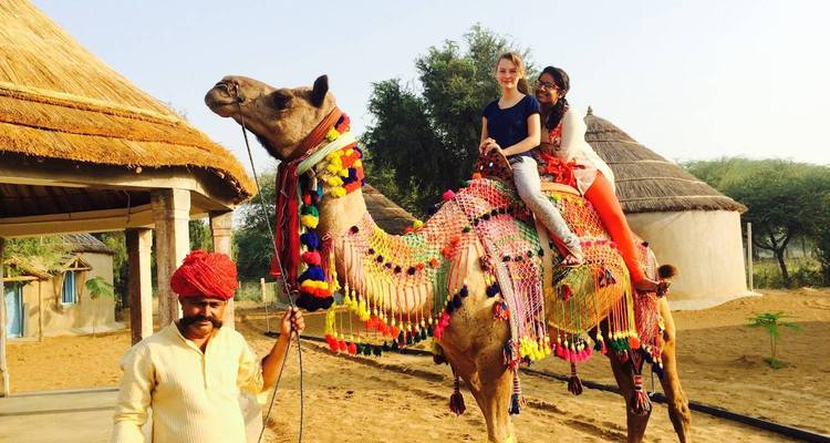 Two travellers ride a brightly decorated camel led by a turbaned handler in a sand-dune village.