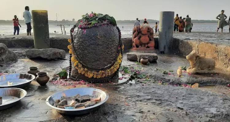 Nahaufnahme eines steinernen Shiva-Lingam, geschmückt mit Blumen und Opfergaben auf einer Gezeitenfelsplattform mit Gläubigen im Hintergrund.