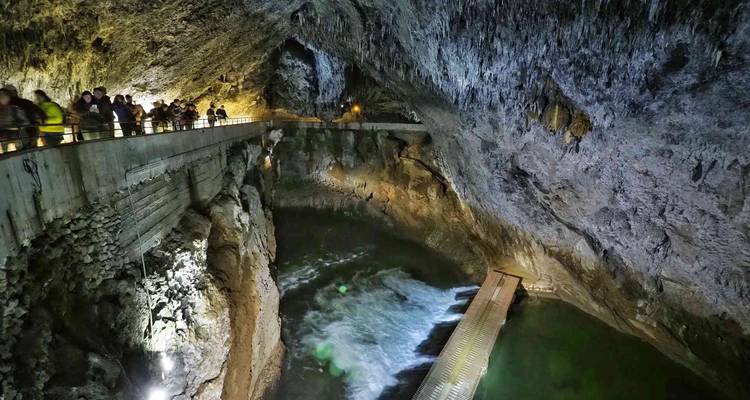 Les touristes marchent le long d'une passerelle en béton éclairée à l'intérieur d'une vaste grotte de calcaire avec une rivière souterraine en dessous.