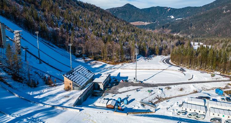 Vue aérienne d'un stade de saut à ski enneigé niché dans les montagnes densément boisées.