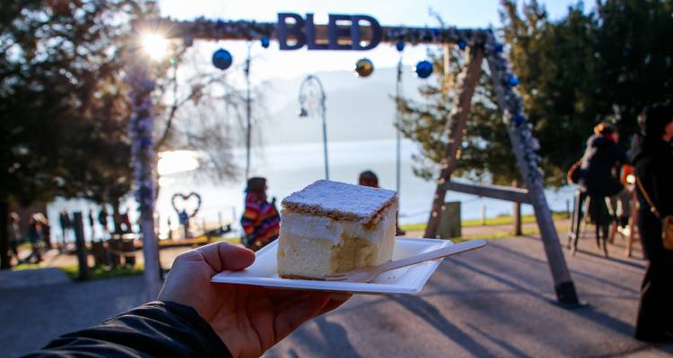 La main tient le célèbre gâteau à la crème de Bled avec une arche festive et le lac en arrière-plan.