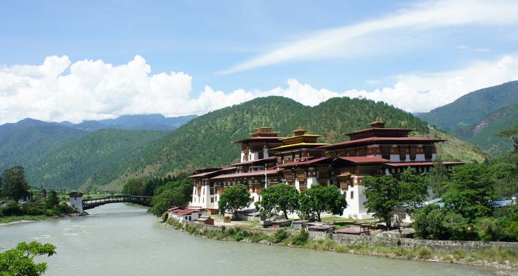 Punakha Dzong elevándose majestuosamente junto a un río ancho con colinas verdes y cielo azul de fondo.