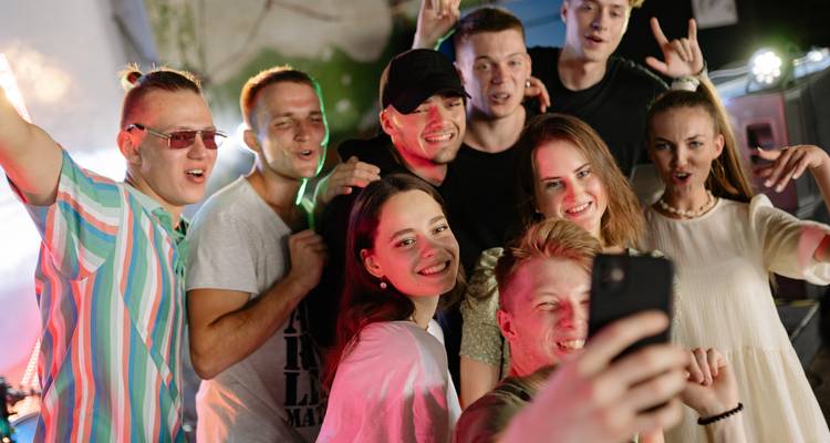 Grupo de jóvenes adultos disfrutando una fiesta en interiores, cantando y tomándose una selfie bajo luces de colores.