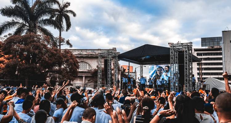 Multitud grande levantando las manos en un concierto de música al aire libre con escenario, pantallas y palmeras.