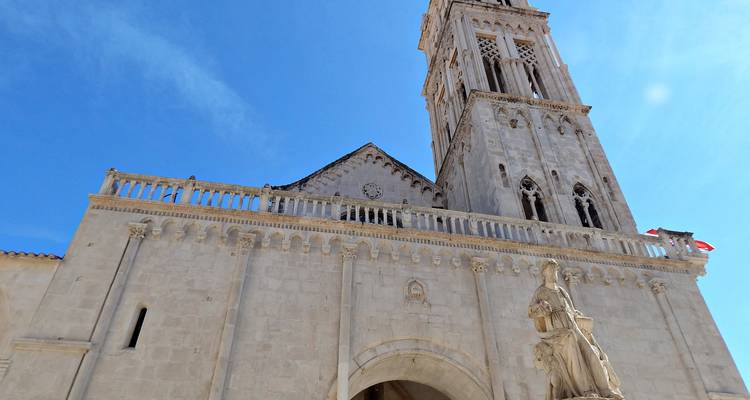 Vista hacia arriba de la fachada de piedra caliza y el campanario de la Catedral de San Lorenzo de Trogir contra un cielo despejado.