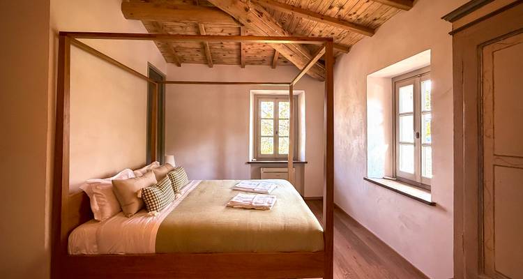 Cozy rustic bedroom with wooden beamed ceiling, four-poster bed, and soft daylight through shuttered windows.