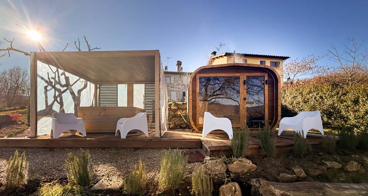 Sunny outdoor wellness area with a wooden sauna hut, pergola lounge and modern white chairs set in a rural Tuscan landscape