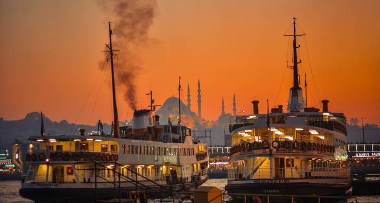 Twin Istanbul ferries at sunset with mosque minarets silhouetted against an orange sky.