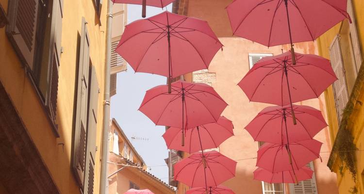 Artistic installation of pink umbrellas suspended above a narrow Mediterranean alley between ochre buildings