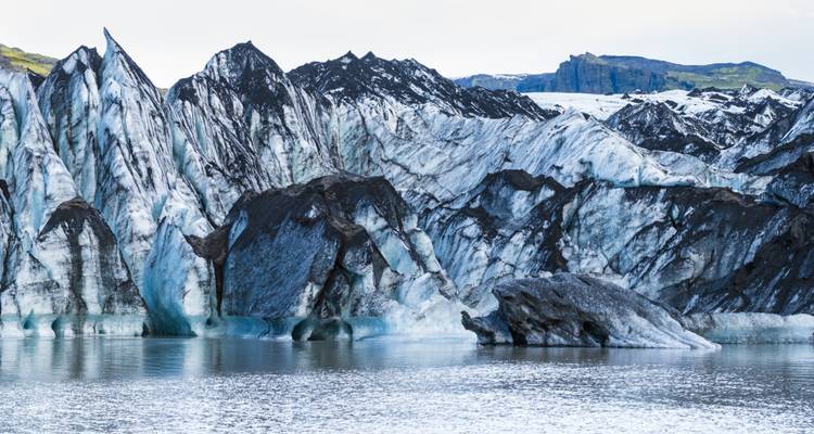 Zerklüftete Gletscher- und schwarze vulkanische Asche, die sich in einer ruhigen Schmelzwasserlagune spiegeln.