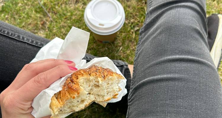 Primer plano de una mano sosteniendo un pastel a medio comer junto a un café para llevar mientras está sentado en el pasto.