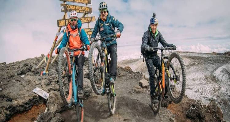 Mountainbikers maken wheelies naast het Uhuru Peak-bordje op de vulkanische bergkam van Kilimanjaro.