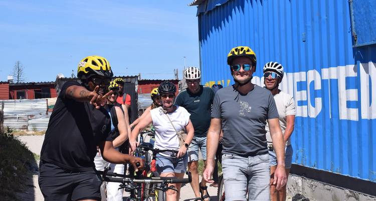 Radfahrer mit Helmen schieben Fahrräder an einer leuchtend blauen Schiffscontainerwand vorbei.