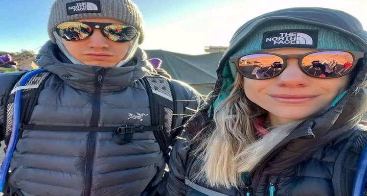 Deux trekkeurs vêtus de doudounes souriants à la caméra pour un selfie lumineux, avec des pics enneigés se reflétant dans leurs lunettes de soleil.