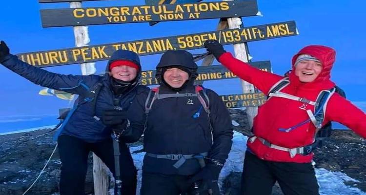 Three climbers in winter gear celebrate beside the Uhuru Peak sign on Mount Kilimanjaro.