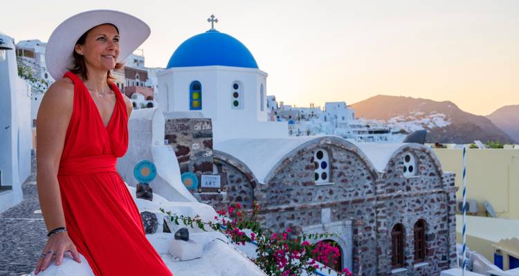 Woman in red dress enjoying Santorini sunset beside blue-domed chapel and pastel rooftops.