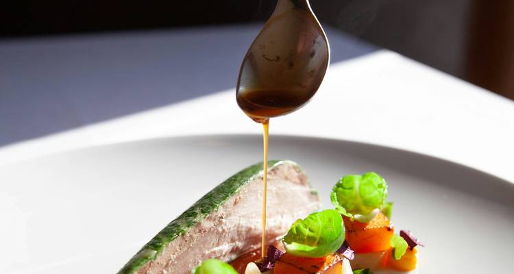 Chef drizzling rich sauce over an artfully plated main course garnished with fresh greens.