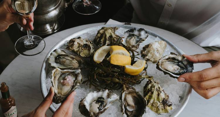 Shared platter of fresh oysters on ice with lemon wedges as diners toast wine glasses.