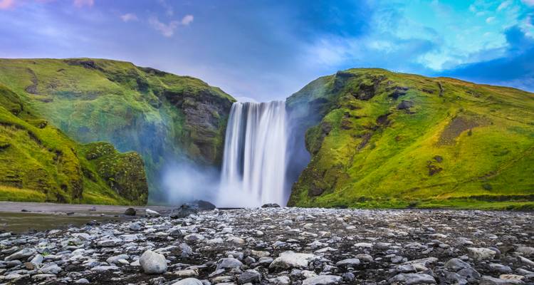 Iconic Skogafoss waterfall cascades over green cliffs with mist rising into a colourful sky.