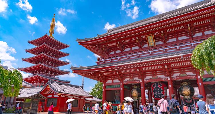 Crowds explore the vibrant red halls and five-story pagoda of Senso-ji under a bright blue sky.
