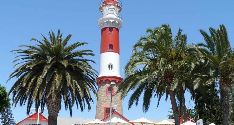 Phare rouge et blanc encadré par de grands palmiers contre un ciel côtier bleu clair.