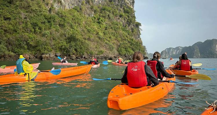 Group of kayakers paddling bright orange boats near a green limestone cliff.