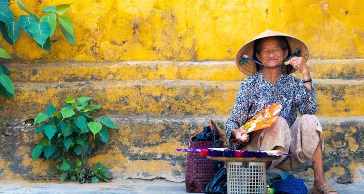 Une femme vietnamienne âgée portant un chapeau conique est assise contre un mur jaune vif et vend des éventails colorés faits à la main.
