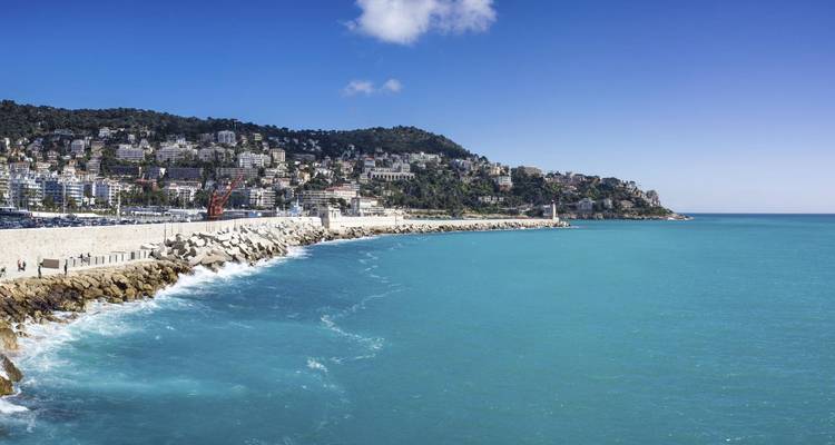 Digue rocheuse et phare gardant la baie turquoise de Nice avec la ville en arrière-plan sur la colline.