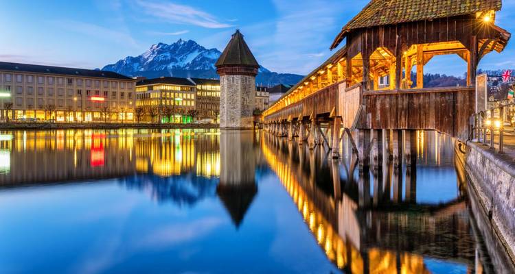 El histórico Puente de la Capilla de madera atraviesa un río tranquilo como un espejo durante la hora azul con el Monte Pilatus recortado contra el cielo de fondo.