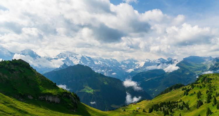 Panorámica alpina expansiva con prados verdes ondulantes, picos rocosos cubiertos de nieve y nubes dramáticas encima.