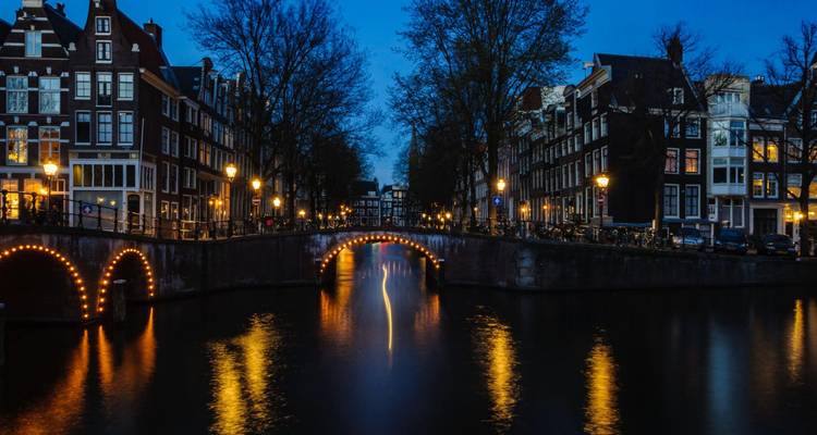 Vista nocturna del canal de Ámsterdam con puentes arqueados iluminados por hileras de luces que se reflejan en el agua oscura.