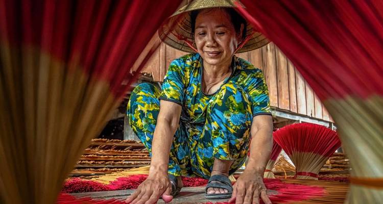 Une artisane vietnamienne vêtue de vêtements colorés arrange des bâtons d'encens à l'intérieur d'un atelier encadré par des rouges éclatants.
