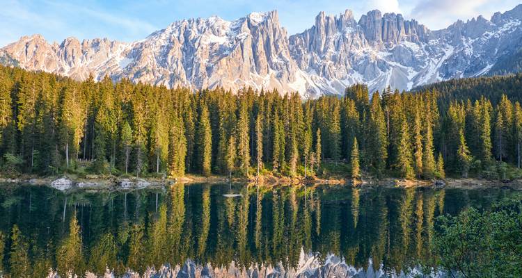 Crystal lake reflecting Dolomite peaks and pine forest at sunrise in the Italian Alps.