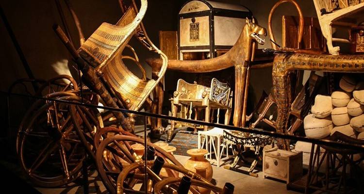 Dimly lit display of pharaoh's chariots, chairs, and treasures from ancient Egypt.