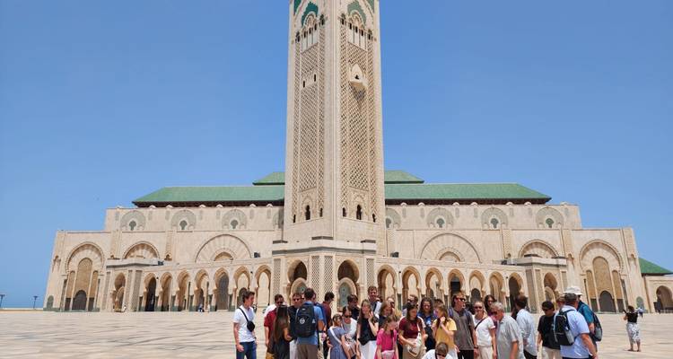 Large tour group stands before the towering minaret and ornate facade of the Hassan II Mosque on a clear day.
