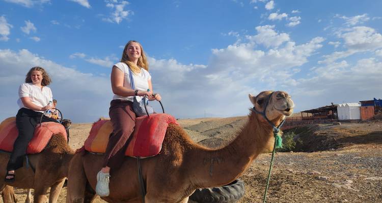 Two women smile while riding camels fitted with red saddles against a dry desert backdrop and bright sky.